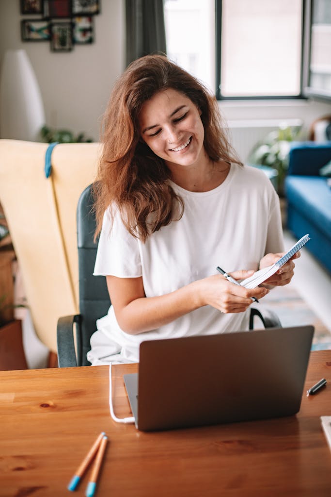 A smiling woman jotting notes while working on a laptop in her home office creating a warm atmosphere.